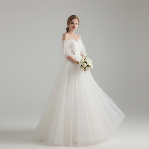 Woman in a white wedding dress holding flowers in a bright room with white curtains.