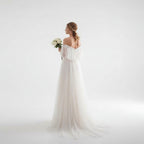 Woman in a white wedding dress holding flowers in a bright room with sheer curtains.