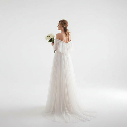 Woman in a white wedding dress holding flowers in a bright room with sheer curtains.