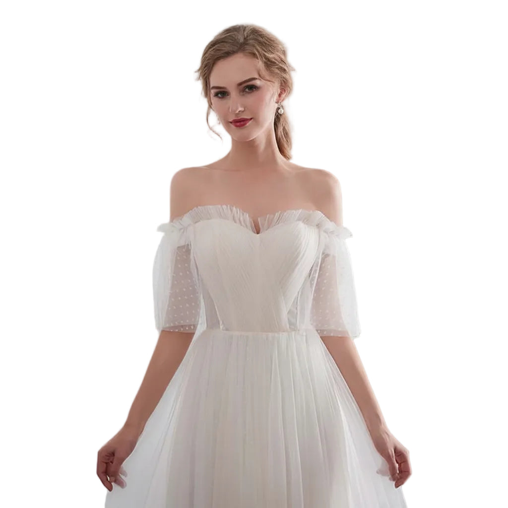 Woman wearing a white off-shoulder dress with sheer sleeves in front of white curtains.