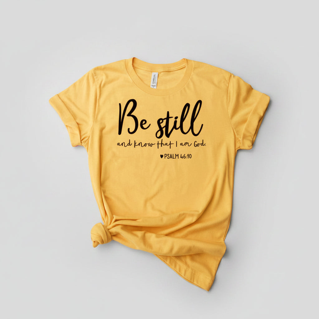 Yellow t-shirt with 'Be still and know that I am God' text, paired with jeans and sneakers on a wooden surface.