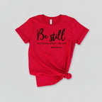 Red t-shirt with 'Be still and know that I am God' text on a wooden surface with shoes and jeans.