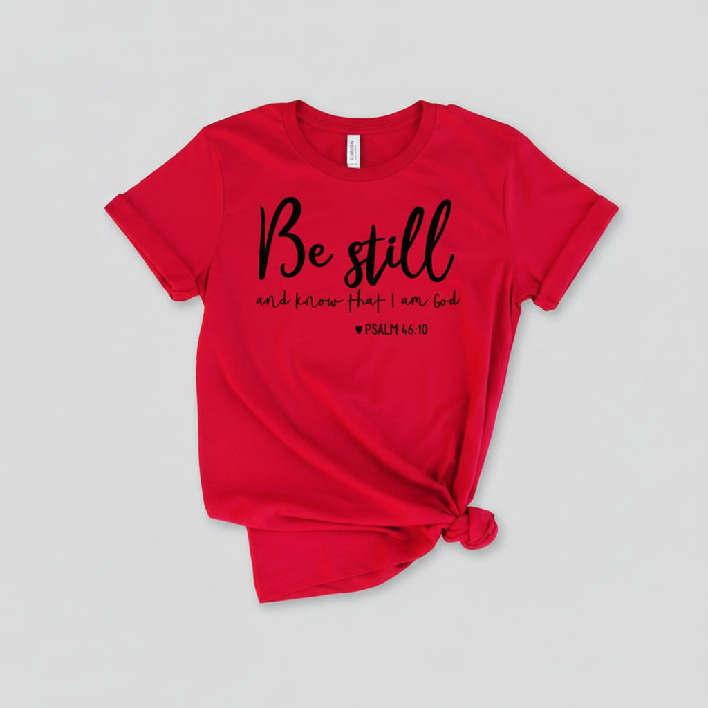 Red t-shirt with 'Be still and know that I am God' text on a wooden surface with shoes and jeans.