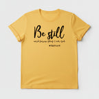 Yellow t-shirt with 'Be still and know that I am God' text, paired with jeans and sneakers on a wooden surface.