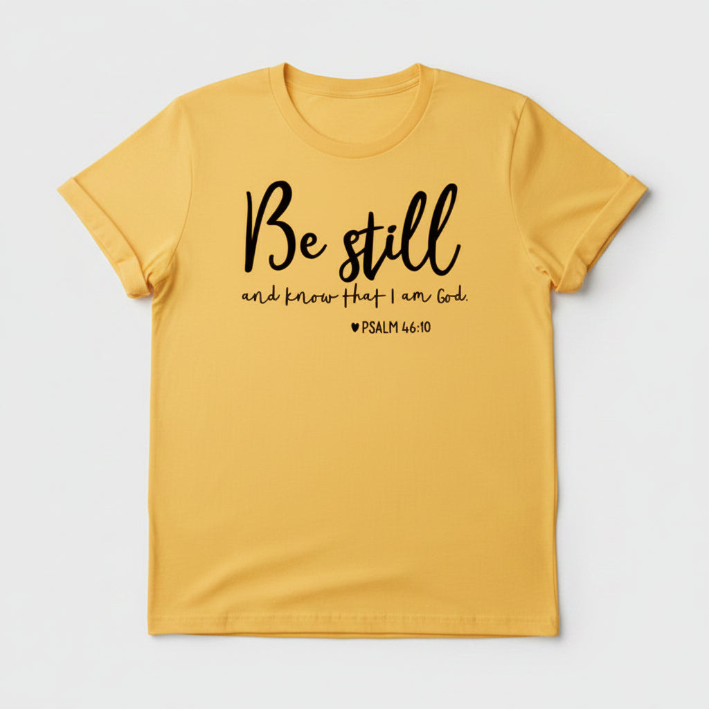Yellow t-shirt with 'Be still and know that I am God' text, paired with jeans and sneakers on a wooden surface.