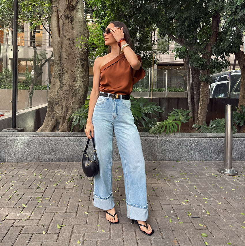 Woman in a rust-colored top and light blue jeans standing outdoors with trees and buildings in the background.