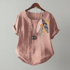 Pink blouse with feather design and buttons on a wooden hanger against a beige wall.