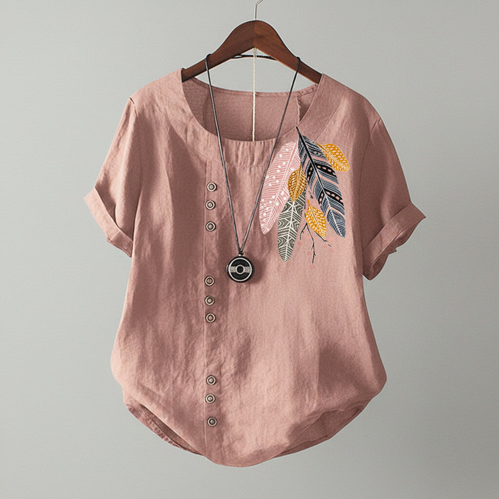Pink blouse with feather design and buttons on a wooden hanger against a beige wall.